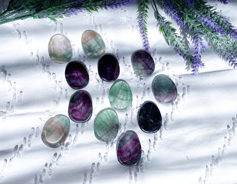 May include: A collection of 12 fluorite crystal stones in various shades of green, purple, and black. The stones are smooth and polished, and they are arranged in a random pattern on a white surface.