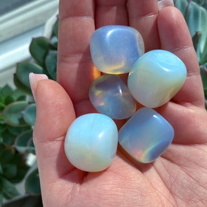 May include: A collection of five smooth, white opal tumbled stones with iridescent rainbow colors. The stones are various sizes and shapes, and are held in a hand.