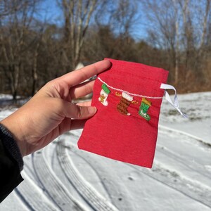 以下が含まれることがあります： 白い紐と刺繍されたクリスマスストッキングが紐からぶら下がっている赤い巾着袋。雪の背景に手の中に持たれている。