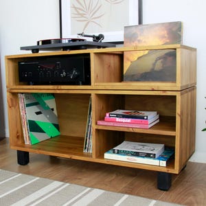 May include: Wooden record player stand with a black turntable, amplifier, and speaker. The stand has compartments for vinyl records and books. The stand is made of light-coloured wood and has black legs.