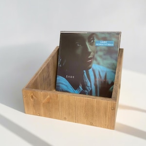 May include: A light brown wooden record holder containing a vinyl record. The record features the album cover of Sade's "The Best of Sade". The album cover shows a portrait of Sade.