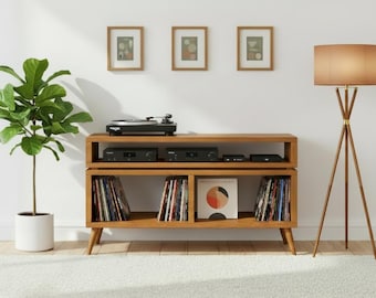 May include: A wooden media console with two open shelves, holding a turntable, audio equipment, and vinyl records. To the left is a potted fiddle leaf fig plant in a white pot, and to the right stands a tripod floor lamp with a tan shade.
