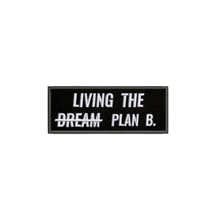 Op de afbeelding: Een zwart rechthoekig embleem met wit geborduurde tekst. De tekst luidt: "LIVING THE DREAM PLAN B." Het woord "DREAM" is doorgestreept. De patch is waarschijnlijk voor kleding of accessoires.