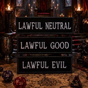Könnte beinhalten: Drei schwarze rechteckige Aufnäher mit weißem Text: "LAWFUL NEUTRAL", "LAWFUL GOOD" und "LAWFUL EVIL". Die Aufnäher werden auf einem Tisch mit Würfeln, einem Schädel und einem Schwert präsentiert, was auf ein Rollenspiel-Thema hindeutet.