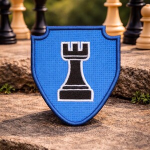 May include: A blue shield-shaped patch featuring a black rook chess piece with a white border. Chess pieces are in the background, indicating a chess theme. The patch is placed on a stone surface.