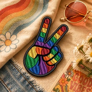 Op de afbeelding: Een kleurrijk geborduurd vredesteken in regenboogkleuren, met een zwarte rand. De patch ligt op een beige stof, naast een oranje getinte zonnebril en een vintage ansichtkaart met de tekst "Good Vibes".