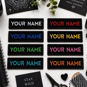 May include: A collection of black rectangular patches embroidered with the text "YOUR NAME" in various colors, including white, red, blue, yellow, green, pink, and orange. The patches are arranged on a white surface, with additional stationery and decorative items.