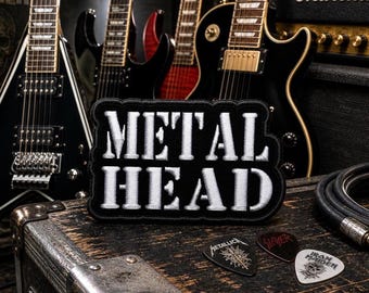 May include: A black and white patch with the text "METAL HEAD" in bold, white letters. The patch is on a wooden surface with electric guitars in the background. Guitar picks with band logos are also visible.