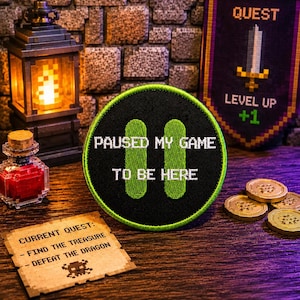 以下が含まれることがあります： 「PAUSED MY GAME TO BE HERE」の文字と2つの緑色のポーズ記号が入ったゲームをテーマにしたパッチ。バナーには「QUEST」と「LEVEL UP +1」と書かれています。小さなカードには「CURRENT QUEST: - FIND THE TREASURE - DEFEAT THE DRAGON」と書かれています。