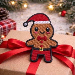 Puede incluir: Un parche de hombre de jengibre con gorro de Papá Noel, pajarita roja y pantalón rojo se coloca en un regalo envuelto con una cinta roja. El parche tiene un cuerpo marrón y un contorno negro. El fondo incluye decoraciones navideñas.
