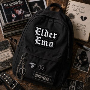 May include: Black backpack with the text "Elder Emo" in white gothic lettering. The backpack has a chain with a heart charm and several pins, including "Misfits" and "blink-182". CDs and a notebook with handwritten text are also visible.