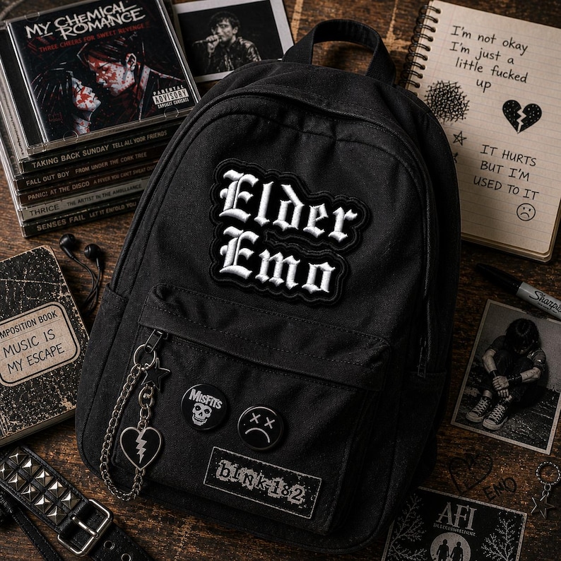 May include: Black backpack with the text "Elder Emo" in white gothic lettering. The backpack has a chain with a heart charm and several pins, including "Misfits" and "blink-182". CDs and a notebook with handwritten text are also visible.