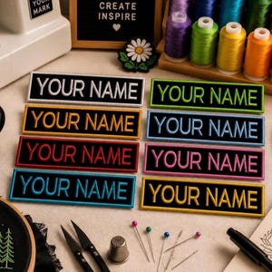 May include: Embroidered patches in various colors, each with the text "YOUR NAME" in white or colored thread. The patches have a black border and are arranged on a light-colored surface. Sewing supplies and spools of thread are also visible.