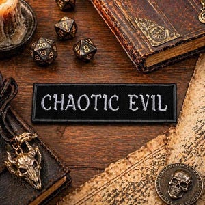 May include: A black patch embroidered with the words "CHAOTIC EVIL" in white. The patch is surrounded by dice, a skull pendant, a vintage book, and a skull coin, all arranged on a wooden surface. The scene evokes a fantasy or role-playing game theme.