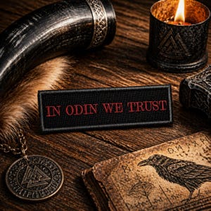 May include: A collection of Viking-themed items on a wooden surface. Includes a black patch with red text reading "IN ODIN WE TRUST", a pendant, a candle, a drinking horn, and a book with a raven illustration. The items are arranged to evoke a sense of Norse mythology.