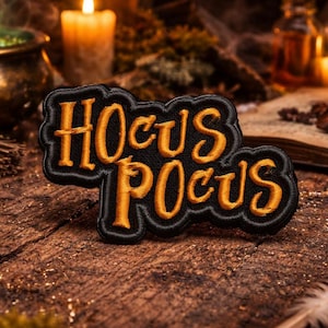 May include: Embroidered patch with the words "Hocus Pocus" in orange script on a black background. The patch is on a rustic wooden surface, with candles and a cauldron in the background, evoking a Halloween or witchy theme.