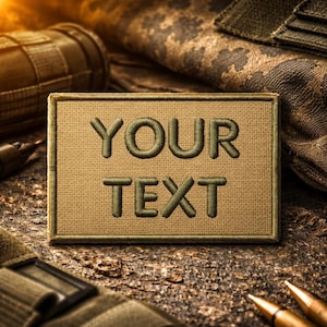 May include: A rectangular, tan patch with the embroidered text "YOUR TEXT" in olive green. The patch is surrounded by a matching olive green border. The background includes military-style gear and camouflage fabric.