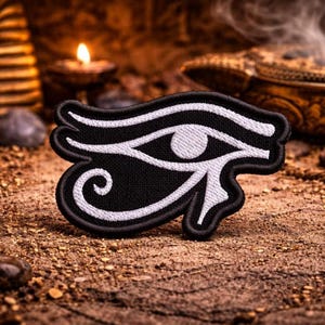 Könnte beinhalten: Schwarz-weißer, gestickter Aufnäher mit dem Horusauge-Symbol. Der Aufnäher hat einen schwarzen Hintergrund mit weißen Details und einen schwarzen Rand. Er liegt auf einer braunen, strukturierten Oberfläche.