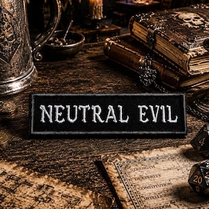 May include: A black rectangular patch with the words "NEUTRAL EVIL" embroidered in white. The patch is displayed on a wooden surface, alongside dice, books, and a decorative metal mug, suggesting a fantasy or role-playing game theme.