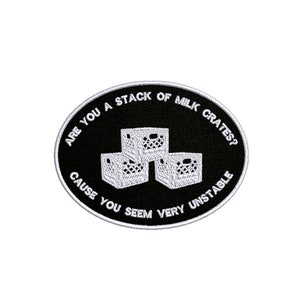 以下が含まれることがあります： 「Are you a stack of milk crates? Cause you seem very unstable.」というテキストと、積み重ねられた3つのミルククレートのグラフィックが描かれた、黒と白の刺繍パッチです。