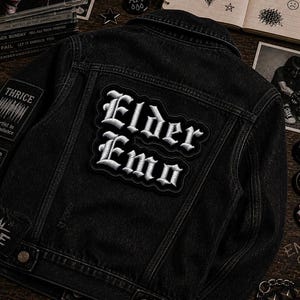 May include: Black denim jacket with patches. The back of the jacket features a large patch with the words "Elder Emo" in white gothic lettering. Additional patches include "THRICE" and other band names. The jacket is on a wooden surface.