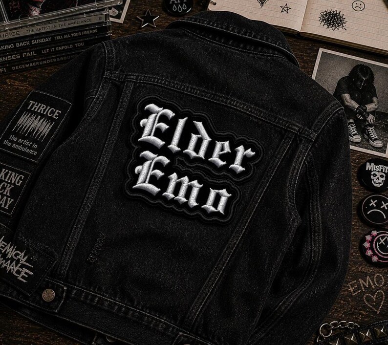 May include: Black denim jacket with patches. The back of the jacket features a large patch with the words "Elder Emo" in white gothic lettering. Additional patches include "THRICE" and other band names. The jacket is on a wooden surface.