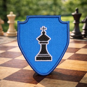 May include: A blue shield-shaped patch featuring a white and black chess king design. The patch is placed on a wooden chessboard, with other chess pieces visible. The patch has a blue embroidered border.