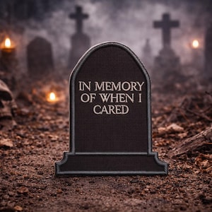 May include: A dark tombstone with the phrase "IN MEMORY OF WHEN I CARED" in white lettering. The scene is set in a graveyard with a skull, candles, and crosses in the background, creating a spooky atmosphere.