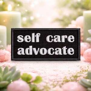 May include: A black patch embroidered with the words "self care advocate" in white. The patch is surrounded by lit candles, succulents, and pink stones, creating a serene and calming aesthetic. The image evokes a sense of wellness.