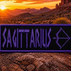 Puede incluir: Un parche rectangular negro con la palabra "SAGITTARIUS" bordada en morado, junto con un símbolo de flecha y arco. El parche está sobre un paisaje desértico con una puesta de sol al fondo.