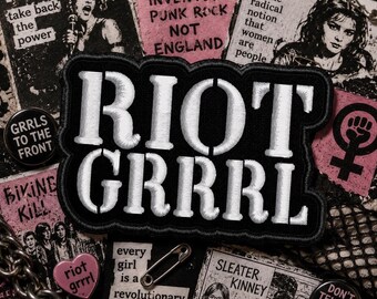 May include: A black and white patch with the words "RIOT GRRRL" in large, white, embroidered letters. The patch is surrounded by other punk rock and feminist themed buttons, pins, and printed materials, including the text "Girls Invented Punk Rock Not England".