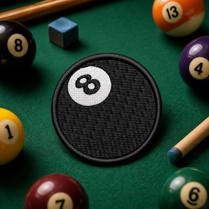 May include: A black, round embroidered patch featuring the number 8 in white, resembling a billiard ball. The patch is surrounded by other billiard balls numbered 1, 4, 6, 7, 8, and 13, a cue stick, and a blue chalk cube, all on a green felt surface.