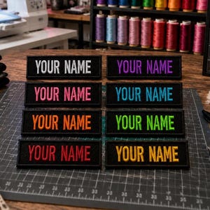 May include: Embroidered patches with the text "YOUR NAME" in various colors, including white, purple, pink, blue, orange, green, and red. The patches are rectangular with a black border and are displayed on a cutting mat.