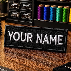 May include: A black rectangular patch with the embroidered text "YOUR NAME" in white. The patch is displayed on a wooden surface, with spools of colourful thread and small storage containers in the background. The patch is likely for customisation.