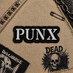May include: A close-up of a tan fabric with several patches and embellishments. The central patch reads "PUNX" in white letters on a black background. Other patches include "DISCHARGE" and a skull graphic. A silver safety pin and chain are also visible.