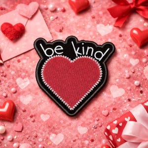 以下が含まれることがあります： 黒い背景に白い文字で「be kind」と刺繍された赤いハート型のパッチ。パッチは白いステッチの縁取りが施されています。画像は、ハートやその他の装飾要素のあるピンク色の背景に設定されています。