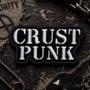Puede incluir: Un parche bordado en blanco y negro con las palabras "CRUST PUNK" en letras negritas y estilizadas. El parche está bordeado en negro y se encuentra sobre un fondo oscuro y texturizado. También se ven un imperdible y una cadena.