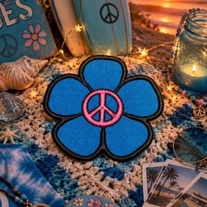 May include: A blue flower-shaped patch with a pink peace sign in the center and a black border. The patch is on a crocheted surface, with other beach-themed items like surfboards and a jar with a candle.