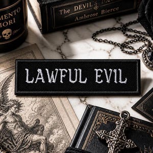 以下が含まれることがあります： 「LAWFUL EVIL」の文字が白で刺繍された黒い長方形のパッチ。本、ネックレス、スカルラベルのボトルなど、ゴシック様式のアイテムが、大理石の表面に配置されています。