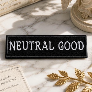 May include: A black rectangular patch with the words "NEUTRAL GOOD" embroidered in white. The patch is displayed on a marble surface, alongside decorative items like a book, a statue, and a gold-coloured chain.