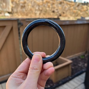 May include: A dark, glossy black bangle bracelet. The circular bracelet is held up against a blurred outdoor background. The bracelet appears to be made of a solid, smooth material.