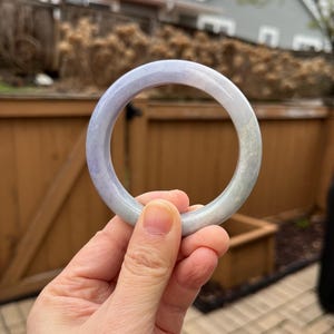 May include: A translucent jade bangle bracelet with a gradient of colors, transitioning from a pale lavender to a soft green. The smooth, polished surface reflects light, showcasing the natural variations in the stone. The bracelet is held against a blurred outdoor background.