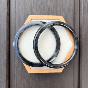May include: Two circular bangle bracelets featuring a dark, mottled black and grey stone-like texture with a polished, glossy finish. The bracelets are positioned overlapping each other, showcasing a smooth, rounded profile and consistent thickness.