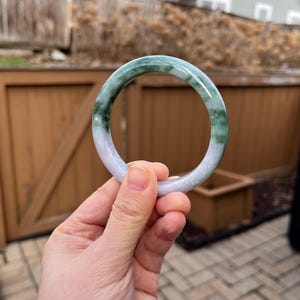 May include: A round jade bangle bracelet with a mix of green and white colors. The jade is translucent, and the bracelet is held in a hand against a blurred outdoor background.