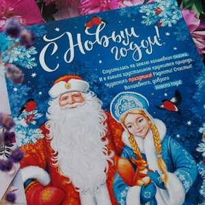 Puede incluir: Una tarjeta de felicitación de Año Nuevo festiva con una ilustración de dibujos animados de Papá Noel y una joven con un vestido azul. La tarjeta tiene un fondo azul con estrellas blancas y el texto "С Новым годом!" que se traduce como "¡Feliz Año Nuevo!" en español.