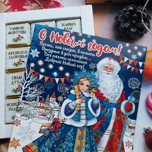 Puede incluir: Una tarjeta de felicitación de Año Nuevo festiva con una ilustración de dibujos animados de Papá Noel y una joven con atuendo tradicional ruso. La tarjeta tiene un fondo azul con estrellas doradas y fuegos artificiales. El texto de la tarjeta dice "S Novym Godom!" que se traduce como "¡Feliz Año Nuevo!" en inglés.