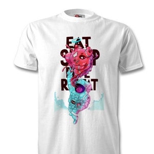 Op de afbeelding: Wit T-shirt met een afbeelding van een astronaut die een roze en blauw wezen vasthoudt met de tekst "Eat Sleep Rocket Repeat" gedrukt op de voorkant.