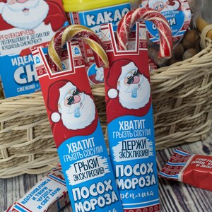 Puede incluir: Dos bastones de caramelo rojo y blanco con envoltorios azules y blancos. Los envoltorios presentan a un Pap&aacute; Noel de dibujos animados con gafas de sol y el texto "Хватит грызть сосули! Грызи эксклюзив! Посох Мороза реплика".