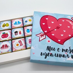 Puede incluir: Una caja azul con un corazón rojo que contiene corazones blancos y una flecha que lo atraviesa. La caja dice "Somos la pareja perfecta" en ruso. La caja está abierta y tiene una bandeja blanca con 12 cuadrados de chocolate con diferentes diseños.