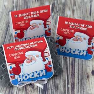 Puede incluir: Tres pares de calcetines con un embalaje festivo rojo y azul con un Pap&aacute; Noel de dibujos animados. El embalaje incluye el texto "NO DEMASIADO PEQUE&Ntilde;O Y NO DEMASIADO ESTRECHO ESTOS SON LOS MEJORES" y "CON EL EFECTO "GUAU" en ruso. Los calcetines son grises con un patr&oacute;n blanco.
